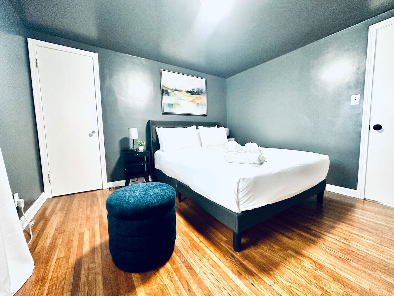 A bedroom with a bed, a nightstand, a wall-mounted painting, and a wooden floor.