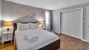 Quarto bem arrumado com uma cama grande, duas mesas de cabeceira com abajures, uma obra de arte na parede e uma porta.