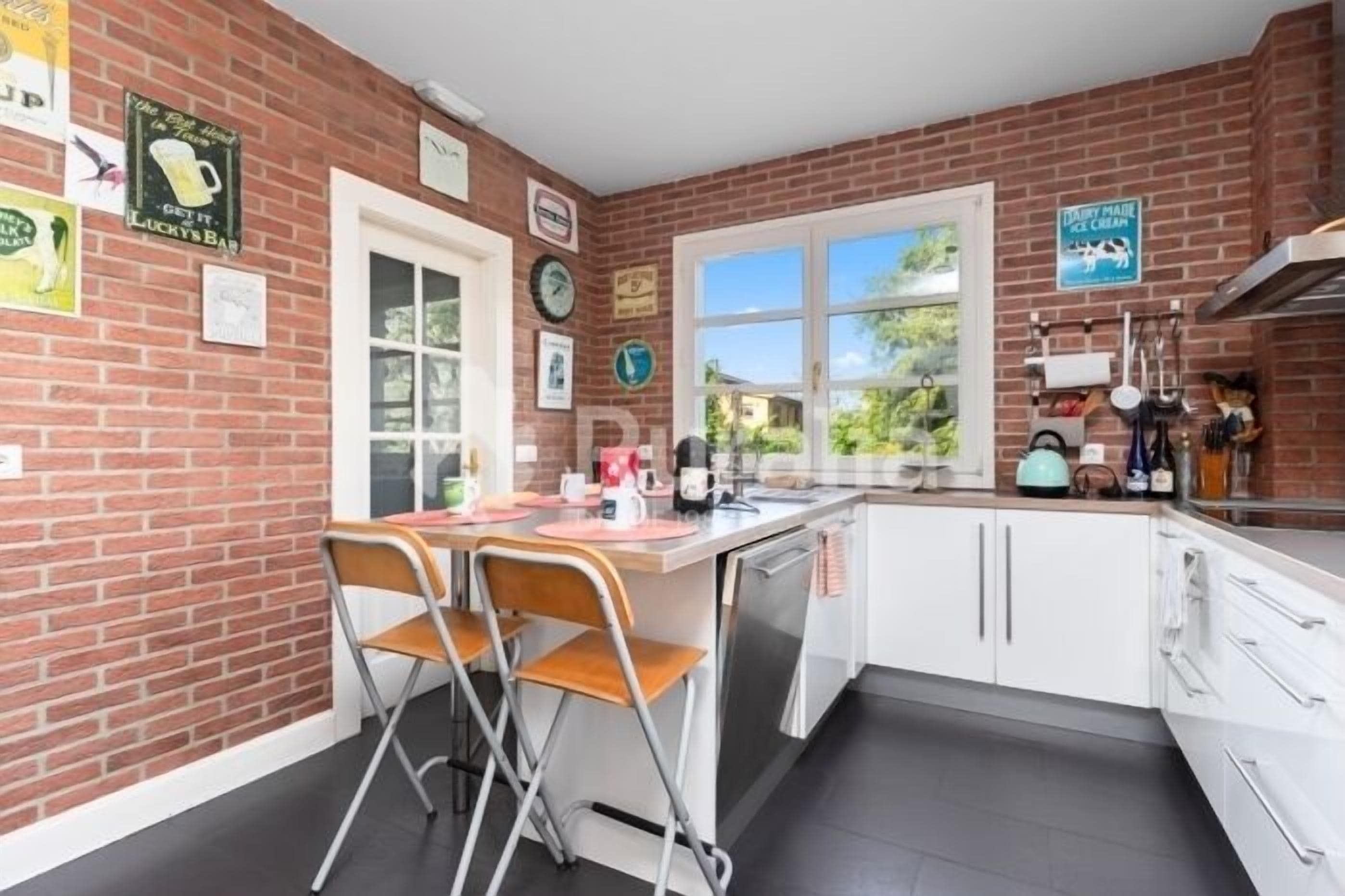 A kitchen with a brick wall, white cabinets, a window, and a dining area with chairs.