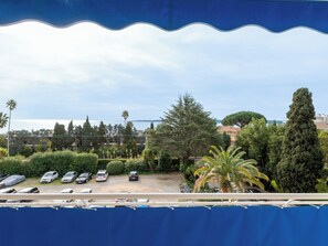 A view of a parking area with several cars, surrounded by greenery and trees, with a body of water and buildings in the background. - 2-Br Sea-View Apartment – East Cannes (Cannes)