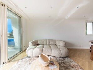 A modern living room with a large sectional sofa, a round coffee table, and a glass sliding door leading to an outdoor area. - 2-Br Sea-View Apartment – East Cannes (Cannes)