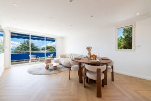 A modern living room with a round dining table, beige sofa, and a large window with a view of greenery and a blue sky. - 2-Br Sea-View Apartment – East Cannes (Cannes)