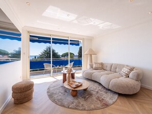 A modern living room with a large window, a round coffee table, a sofa, and a small ottoman. - 2-Br Sea-View Apartment – East Cannes (Cannes)
