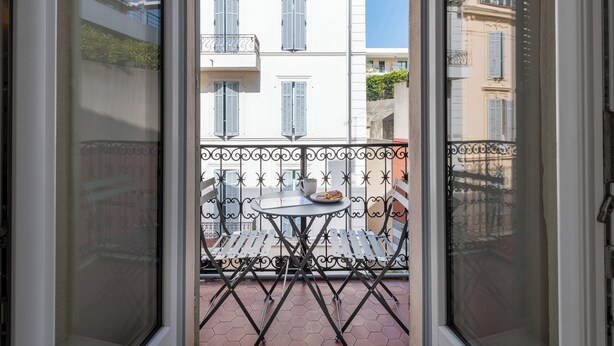 Un balcon avec une table et des chaises, donnant sur un immeuble aux murs blancs et aux volets bleus.