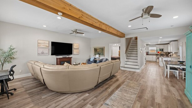 A spacious living room with a large beige sectional sofa, a flat-screen TV, and a ceiling fan. A staircase leads to a kitchen area with white cabinets and a dining table. - Sunflower Yellow Coastal Escape (Galveston)