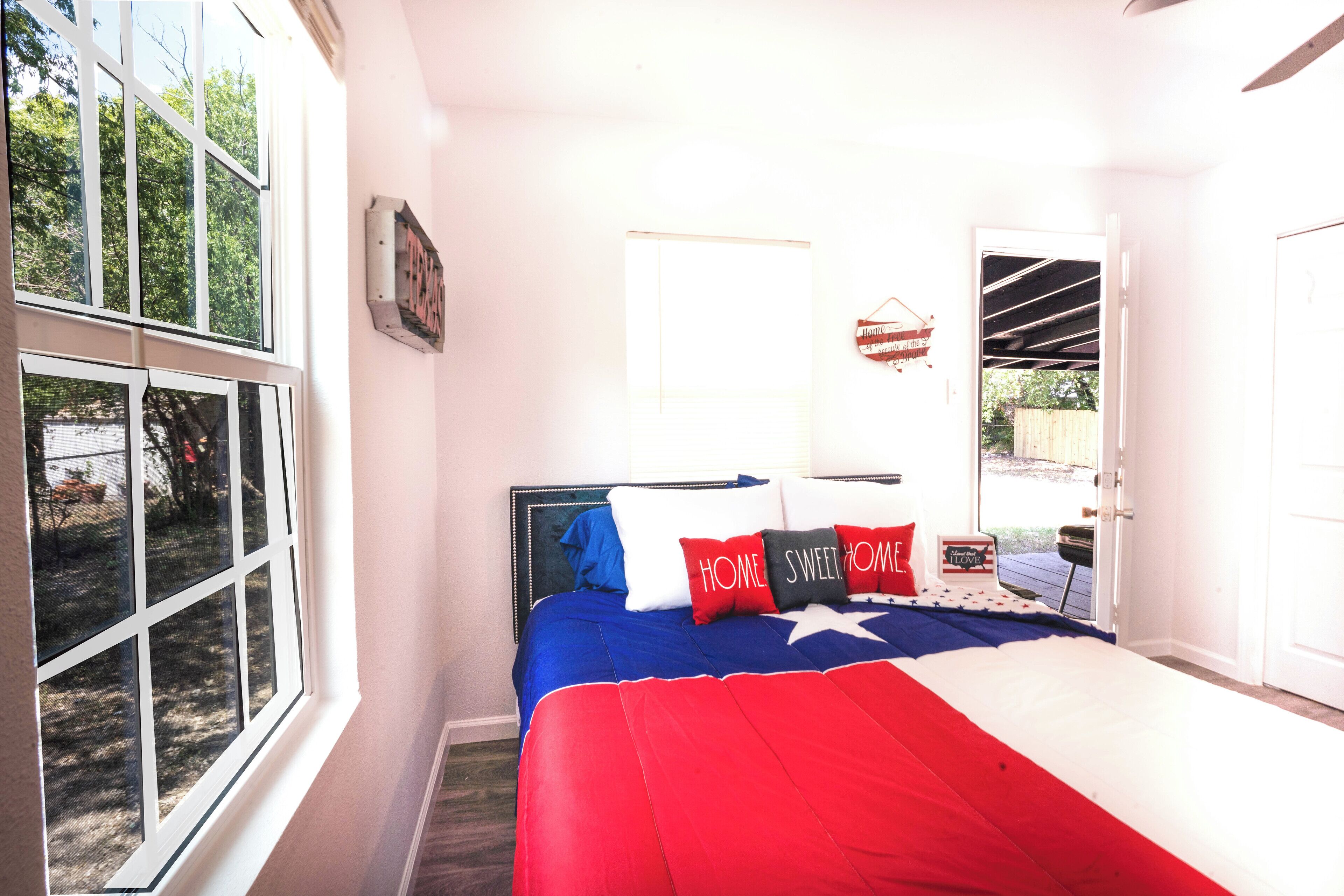 Ein Schlafzimmer mit einem großen Fenster, einem Bett mit einer roten, weißen und blauen Tagesdecke sowie einer Tür, die auf eine Terrasse führt.