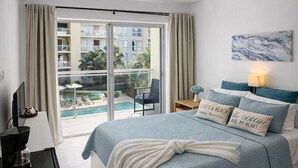 Ein Schlafzimmer mit Bett, Deckenventilator, Fernseher, Kaffeemaschine und Blick auf Pool und Gebäude im Freien.