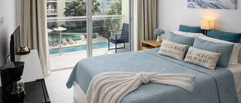 A bedroom with a bed, a ceiling fan, a TV, a coffee maker, and a view of a pool and buildings outside.