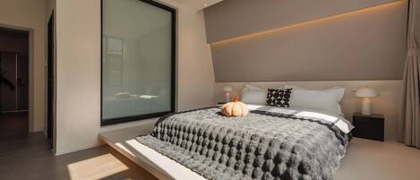 A modern bedroom with a large bed, a window, and a bedside table with a lamp.