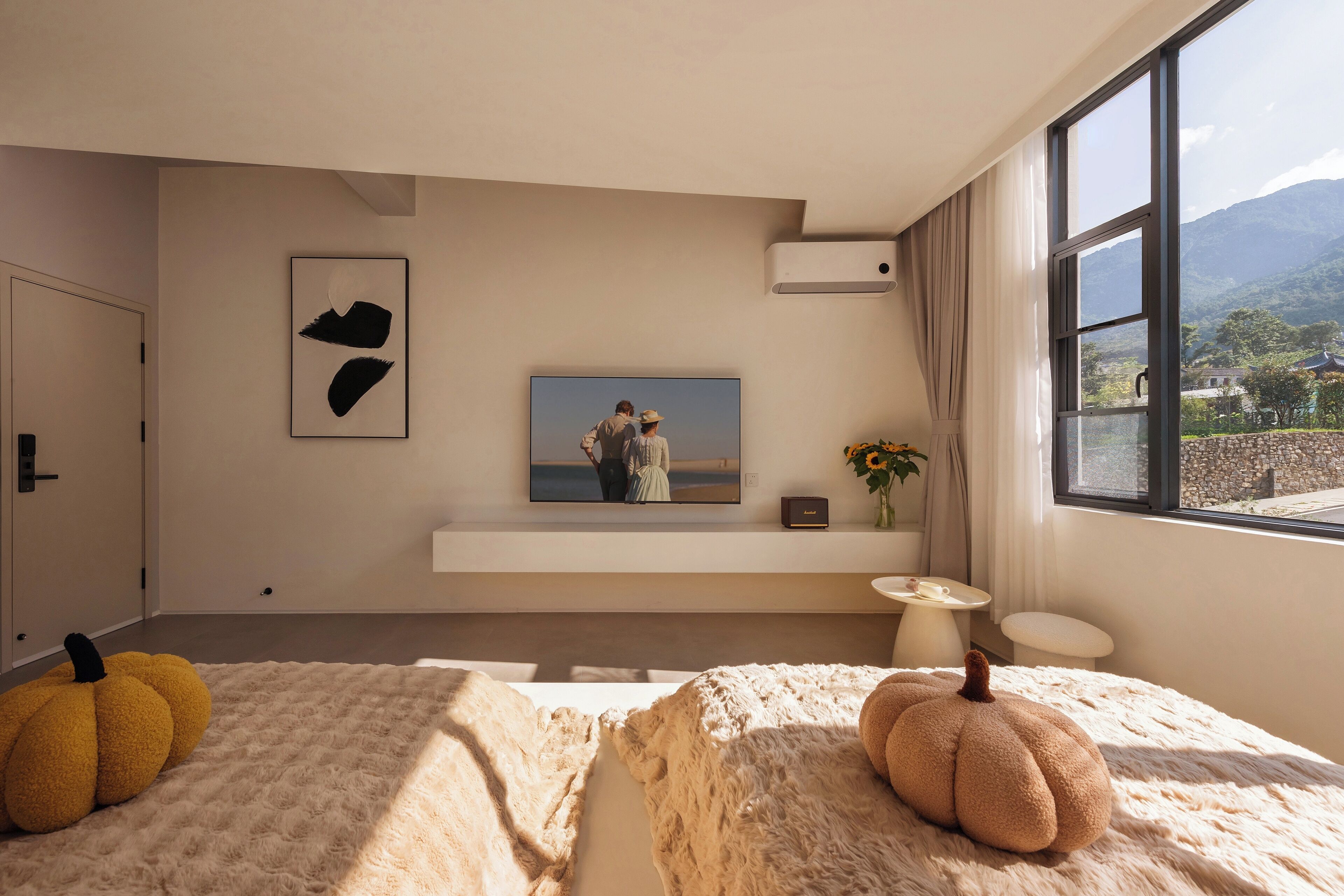 Un dormitorio moderno con un ventanal grande, un televisor de pantalla plana montado en la pared y una cama acogedora con dos calabazas decorativas.