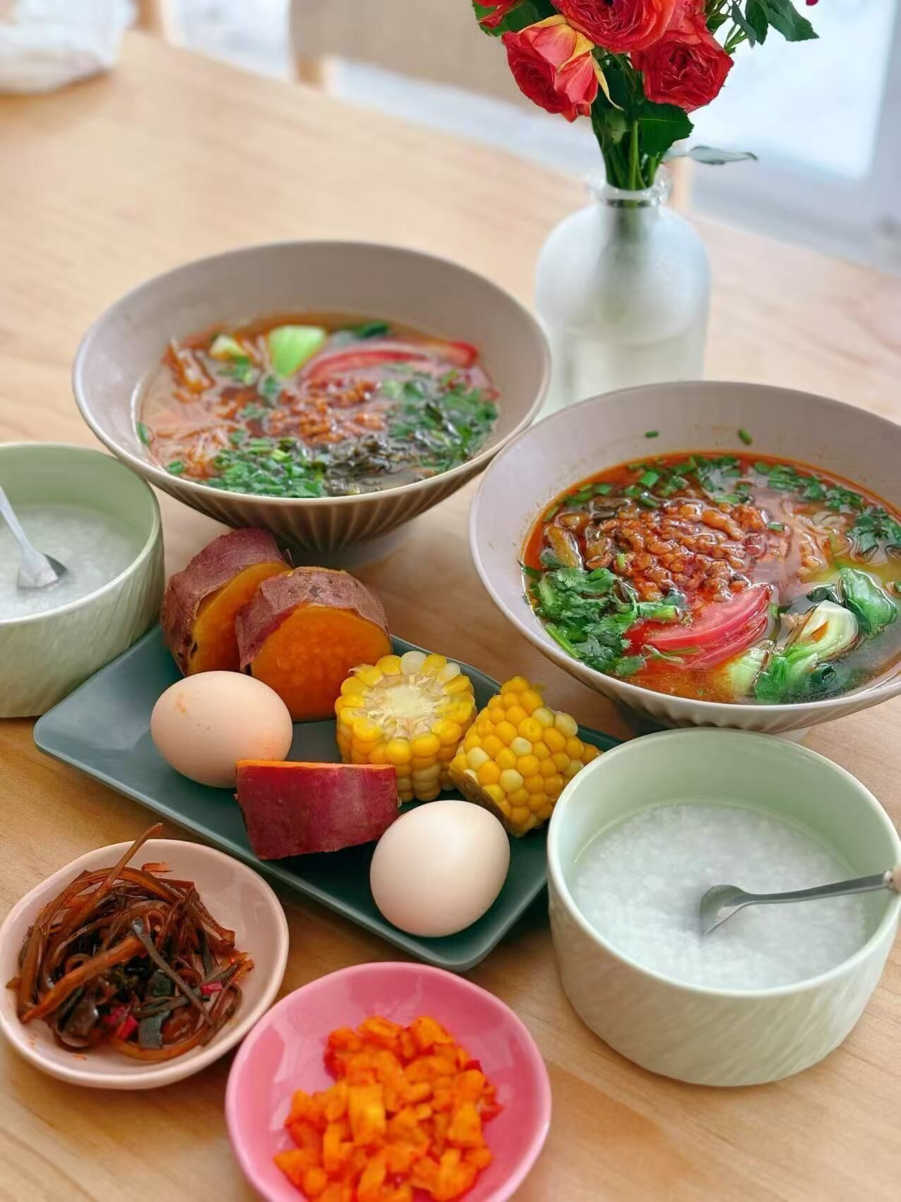 Una mesa con varios platos, entre ellos, tazones de sopa, un plato con maíz, huevos y verduras, y un florero con flores.