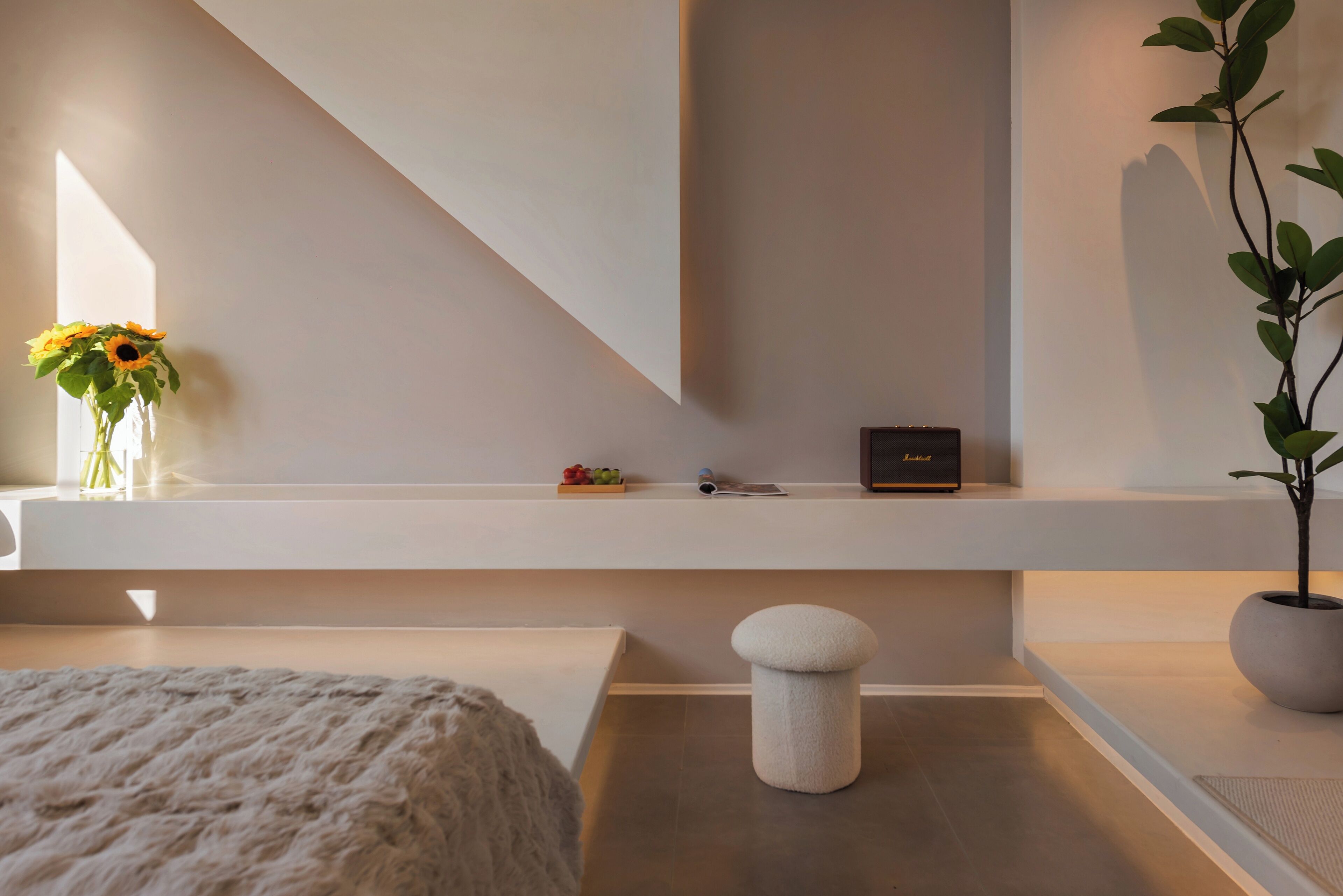 A modern hotel room with a minimalist design, featuring a large window, a white sofa, a small round stool, a potted plant, and a shelf with a vase of flowers and a few items.