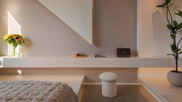 A modern hotel room with a minimalist design, featuring a large window, a white sofa, a small round stool, a potted plant, and a shelf with a vase of flowers and a few items.