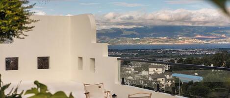 A balcony with two wicker chairs and a small table, overlooking a coastal cityscape with mountains and a body of water.