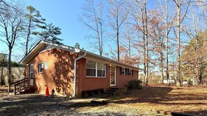 A single-story brick house with a front porch, a small attached garage, and a yard with trees and a wooden deck. - Duke Stayhouse (Durham)