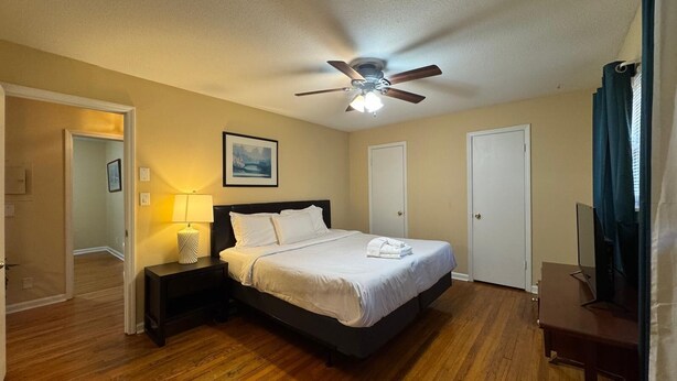 A hotel room with a bed, a ceiling fan, a nightstand with a lamp, and a painting on the wall. - Duke Stayhouse (Durham)