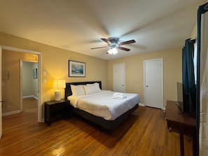 Habitación de hotel con cama, ventilador de techo, mesita de noche con lámpara y un cuadro en la pared.