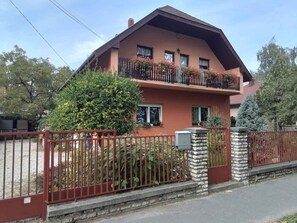 A two-story house with a red facade, a balcony with flowers, and a stone pillar. - Holiday apartment near the beach with Wi-Fi and air conditioning (Siófok)