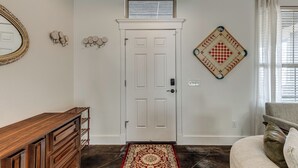 Un couloir avec une porte blanche, une console en bois, un tapis à motifs, un jeu d’échecs mural et un luminaire suspendu.