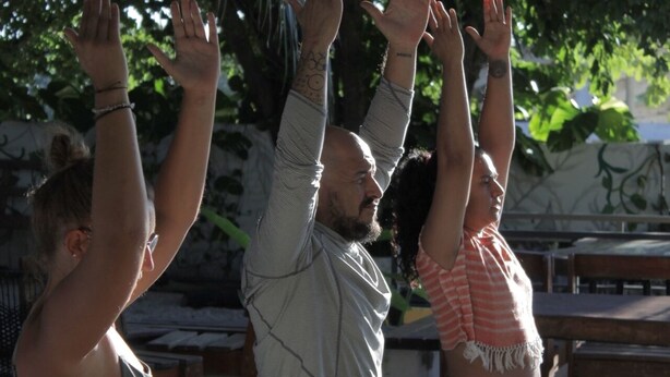 Tre personer utövar yoga utomhus, på en plats med ett tak och trämöbler.