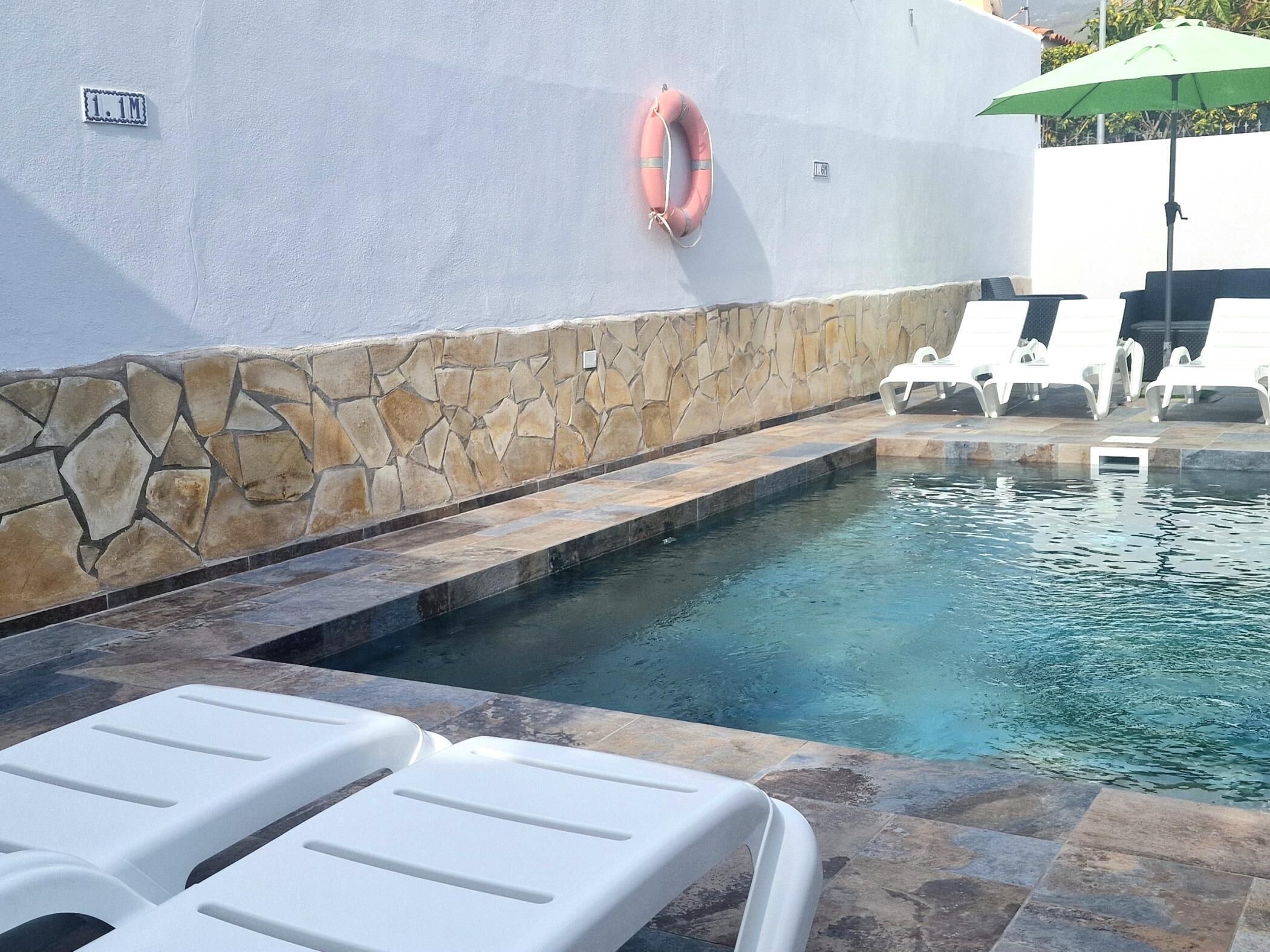 Une piscine aux bords recouverts de carrelage en pierre, des chaises longues blanches et une bouée de sauvetage fixée au mur.