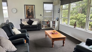 A living room with a black leather sofa, a wooden coffee table, a bookshelf, and a large window with a view of trees.