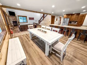 A spacious living room with a large wooden dining table, a fireplace, and a kitchen area with wooden cabinets.