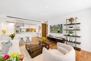 A modern living room with a glass coffee table, a comfortable armchair, a TV mounted on the wall, and a kitchen area with white cabinets and a stainless steel range hood. - Exclusive Fitzrovia 3-bed Concierge Central AC (London)