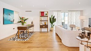 Una sala de estar moderna con piso de madera, un sofá blanco, una mesa de centro, una mesa de comedor con sillas, una estantería y un amplio ventanal con cortinas.