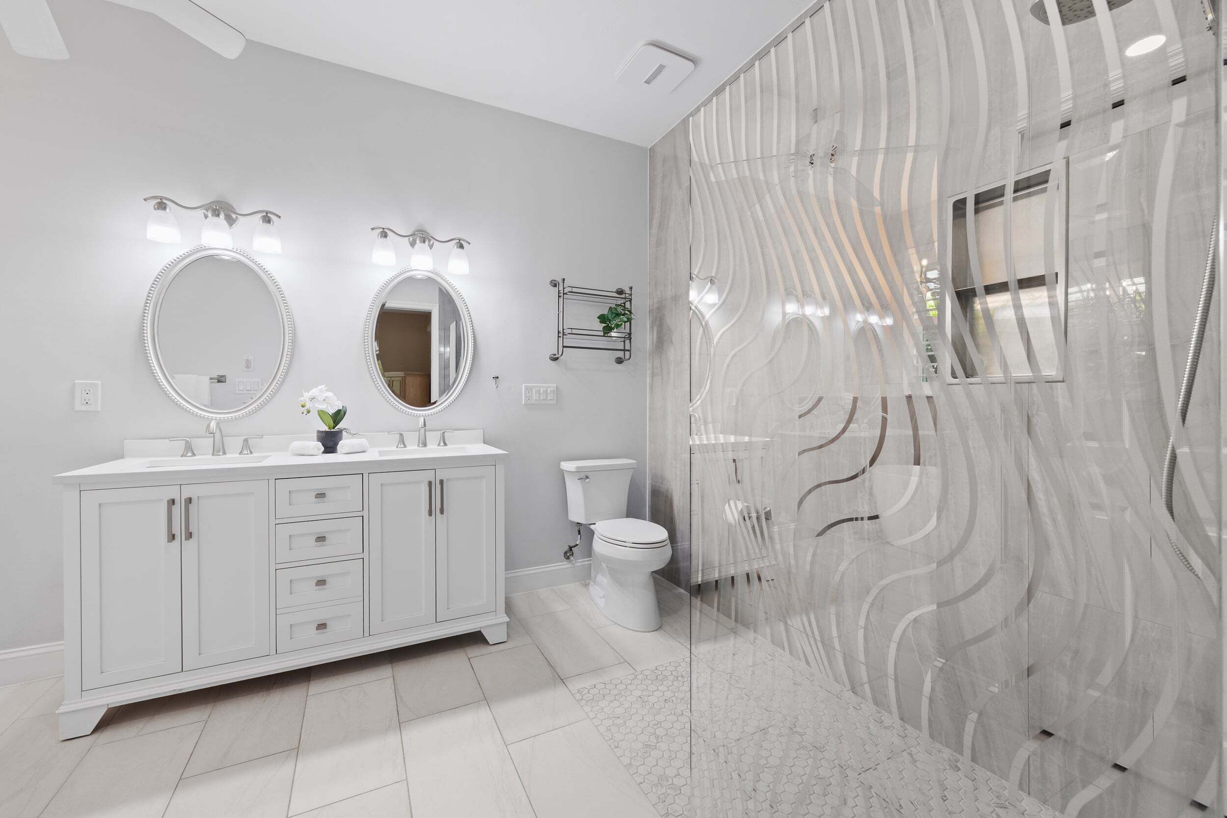 Ein modernes Badezimmer mit einem weißen Waschtisch, zwei Spiegeln, einer Toilette und einer Duschkabine mit wellenförmigem Fliesendesign.