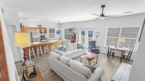 En rummelig opholdsstue med sofa, sofabord og et spisebord med stole. Et køkken med køleskab og skabe kan ses i baggrunden.