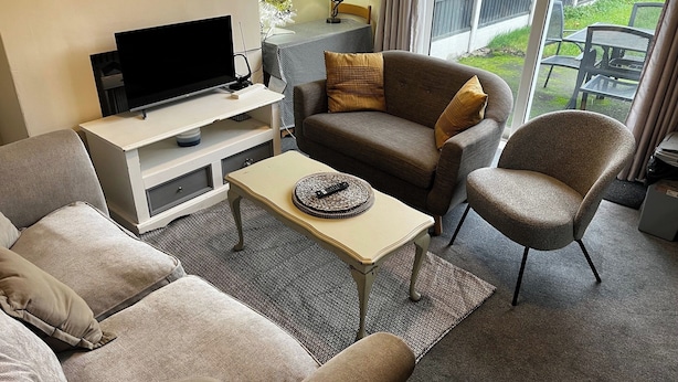 Ett vardagsrum med en soffa, en fåtölj och ett soffbord, en platt-TV och ett fönster med utsikt över omgivningarna.