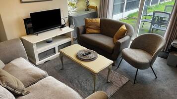 A living room with a sofa, armchair, and coffee table, a flat-screen TV, and a window with a view of the outdoors.