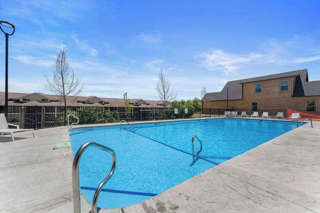 A rectangular swimming pool with metal ladders on one side, surrounded by a concrete deck. There are lounge chairs and a building with a sloped roof in the background.