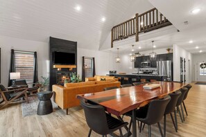 A spacious living room with a large wooden dining table, comfortable seating, a fireplace, and a kitchen area with modern appliances.