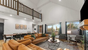 A spacious living area with a high ceiling, a wooden staircase, and a dining area with modern furniture. - Fire Pit! Hot Tub! Stunning Views! Two Fireplaces! (Branson)
