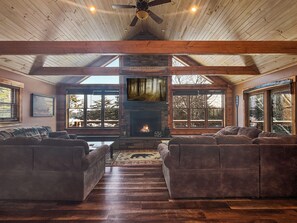 A spacious living room with a stone fireplace, wooden beams, and large windows offering a view of the outdoors.