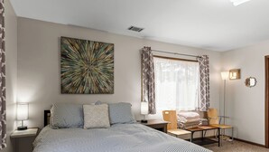 Hotellihuone, jossa on suuri sänky, työpöytä tuolilla, lattiavalaisin, ikkuna verhoilla ja seinälle kiinnitetty taideteos.