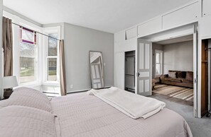 A bedroom with a bed, a mirror, and an open door leading to a living area with a sofa.