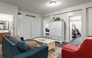 Un salon comprenant un canapé, une table basse, un fauteuil rouge, une armoire et un lit recouvert d’une couverture bleue.