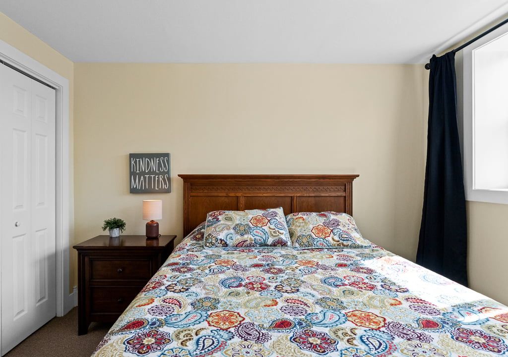 A bedroom with a bed, a nightstand, a wall sign, and a window with curtains.