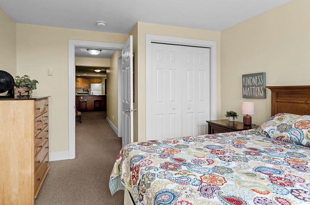 A bedroom with a bed, a dresser, a door, and a kitchen in the background.