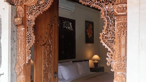 An intricately carved wooden door leading into a room with a bed, a nightstand, and a lamp. - Pande Permai Guest House (Ubud)