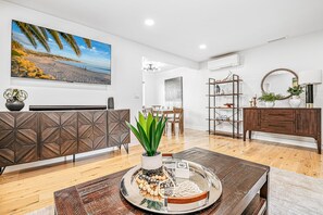 Ein modernes Wohnzimmer mit Couchtisch aus Holz, einem Essbereich mit einer Chandelieber und einem großen Fernseher, der eine Strandansicht zeigt.