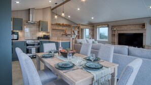 Una cocina moderna con electrodomésticos integrados, un comedor con mesa de madera y sillas tapizadas en gris, y una sala de estar con sofá y chimenea.