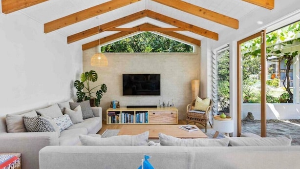 Ein modernes Wohnzimmer mit einer großen Couch, einem Couchtisch, einem an einer weißen Wand montierten Fernseher und Holzbalken an der Decke.
