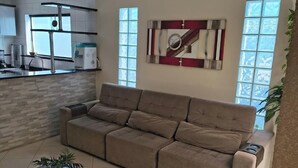 Een moderne woonkamer met een beige bank, een glazen salontafel en een keukenhoek met een raam en een decoratief wandobject.