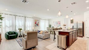 Ein modernes Wohnzimmer mit Sofa, Sessel, Essbereich, Kücheninsel und Kühlschrank.