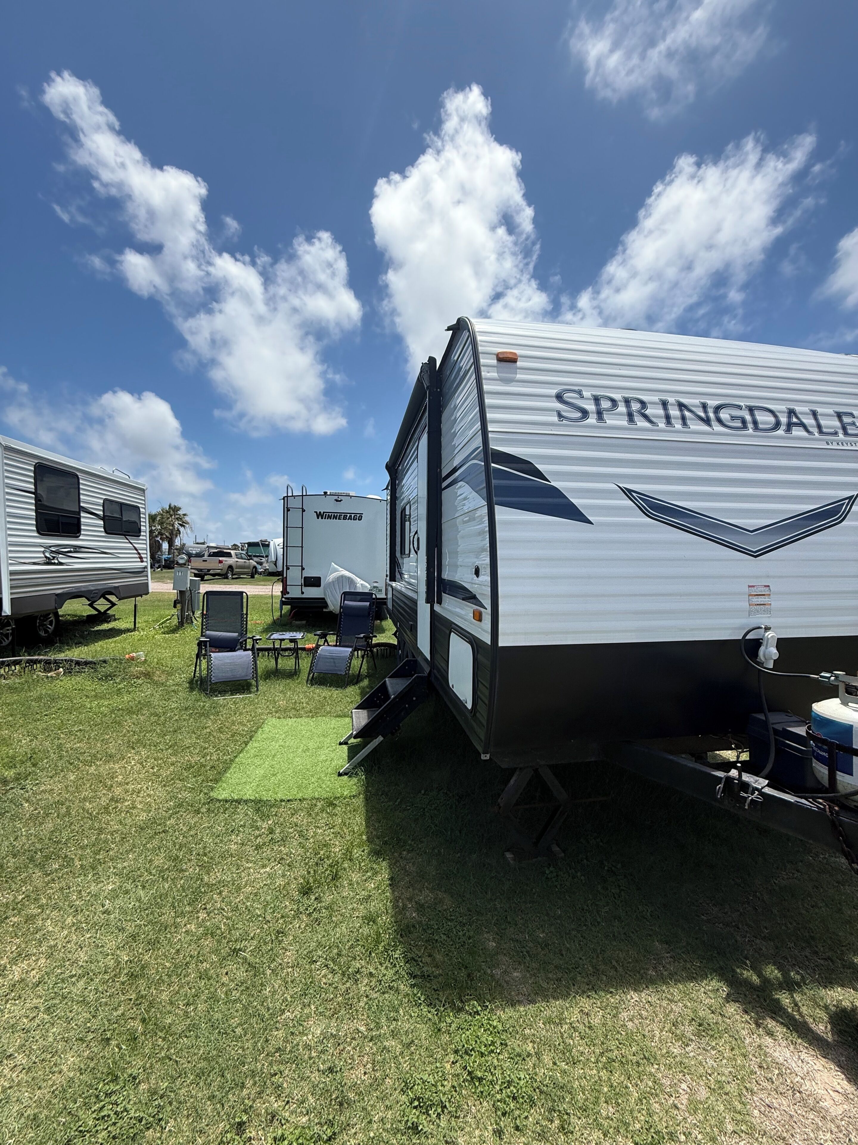 Una caravana recreativa con las marcas SPRINGDALE y WINNEBAGO visibles, estacionada sobre césped con otras caravanas al fondo.