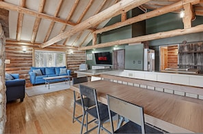 A log cabin interior with a wooden dining table, blue sofa, and a TV mounted on the wall. - Pigeon Retreat Firepit Playset Dock (Clintonville)